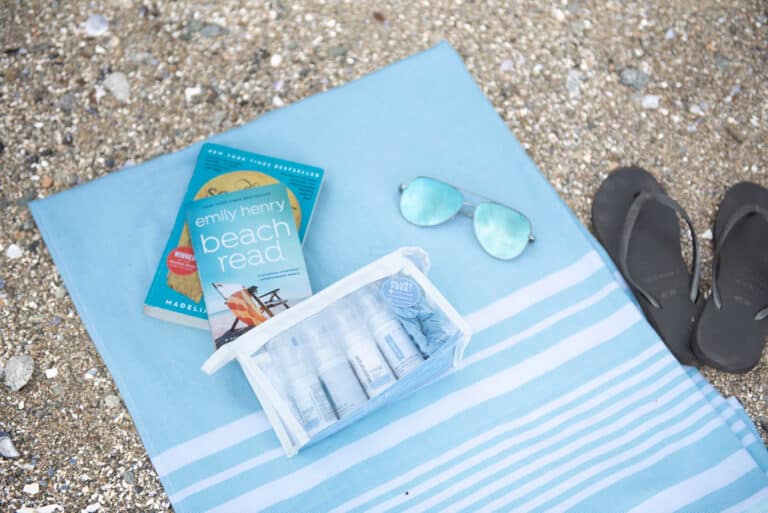 A flat lay at the beach of a blue blanket, flip flops, sunglasses, books and skin care products for sunburn relief and protection